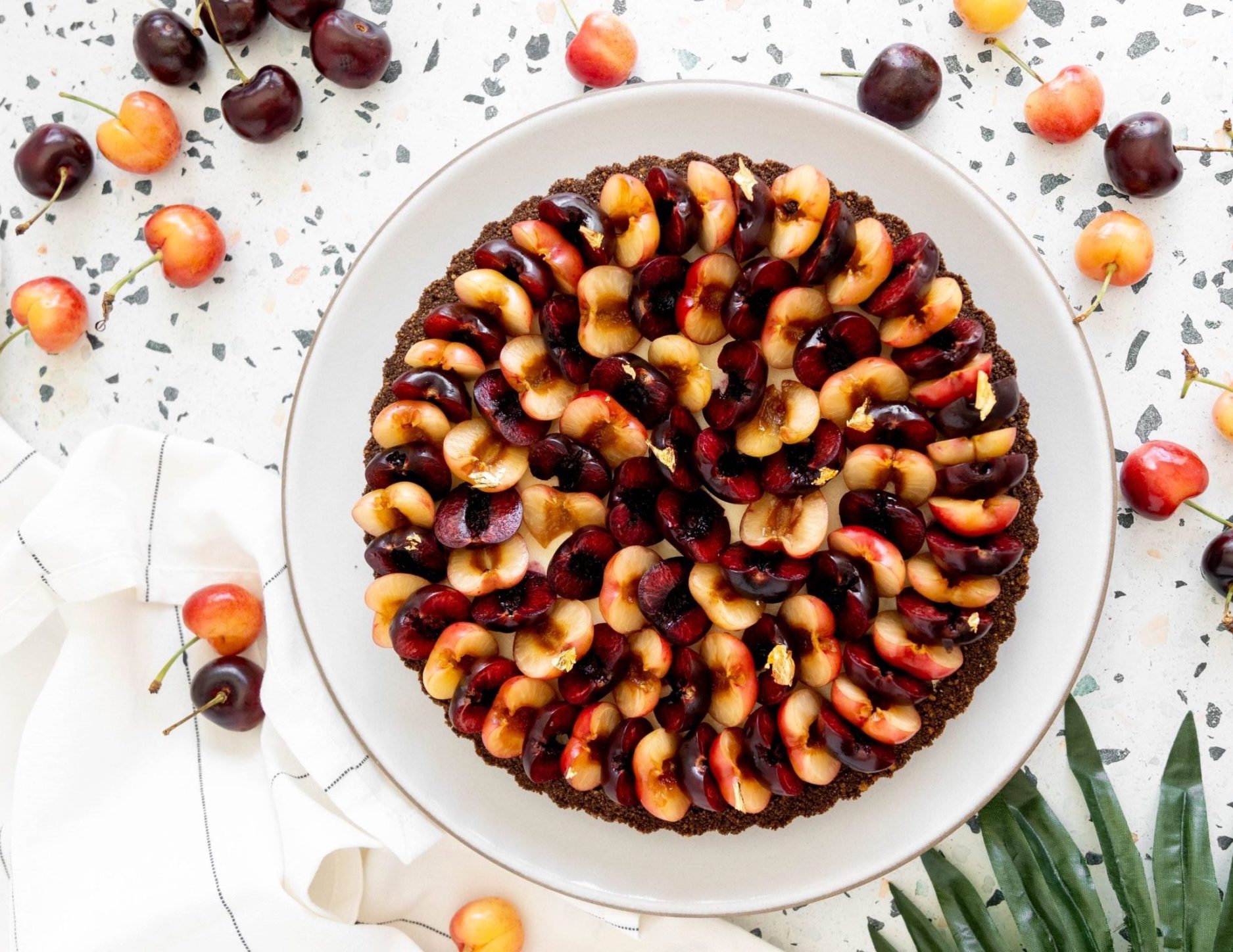 Round tart topped with rows of halved dark red and yellow-orange cherries, garnished with gold leaf, surrounded by whole c...