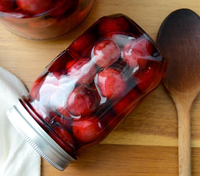 Glass jar filled with red cherries preserved in liquid, lying on a wooden surface next to a wooden spoon.